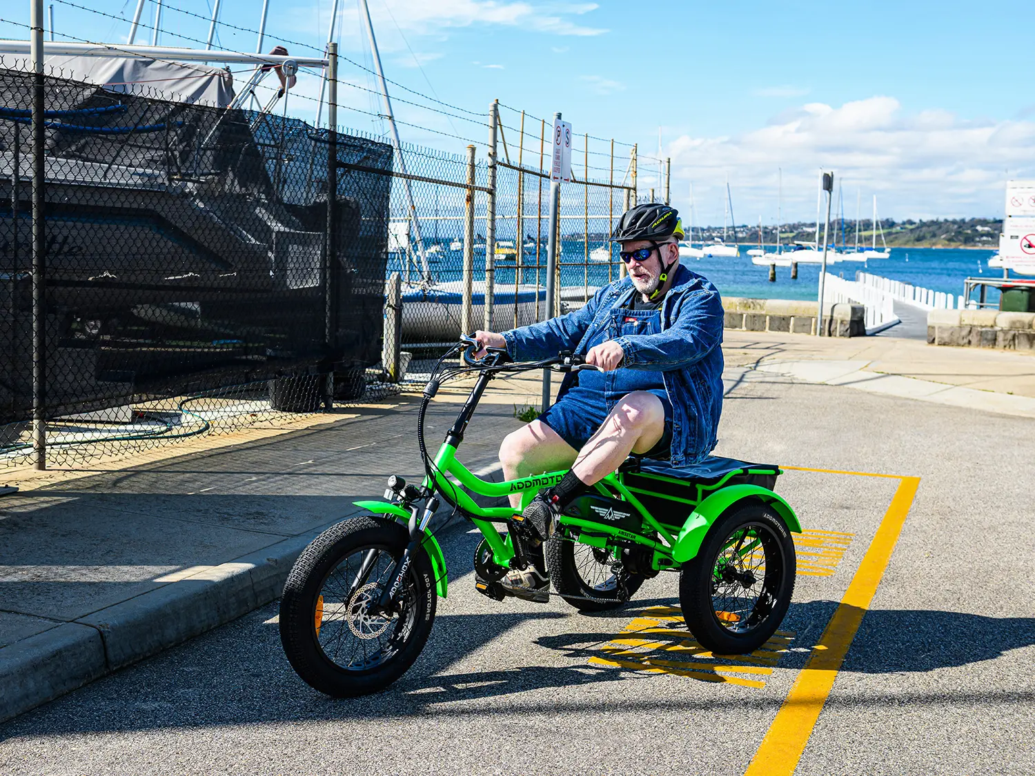 Older man wearing a helmet riding the M-360 electric tricycle along a harbor with sailboats in the background. The tricycle features fat tires and a sturdy frame, designed for stability and comfort.